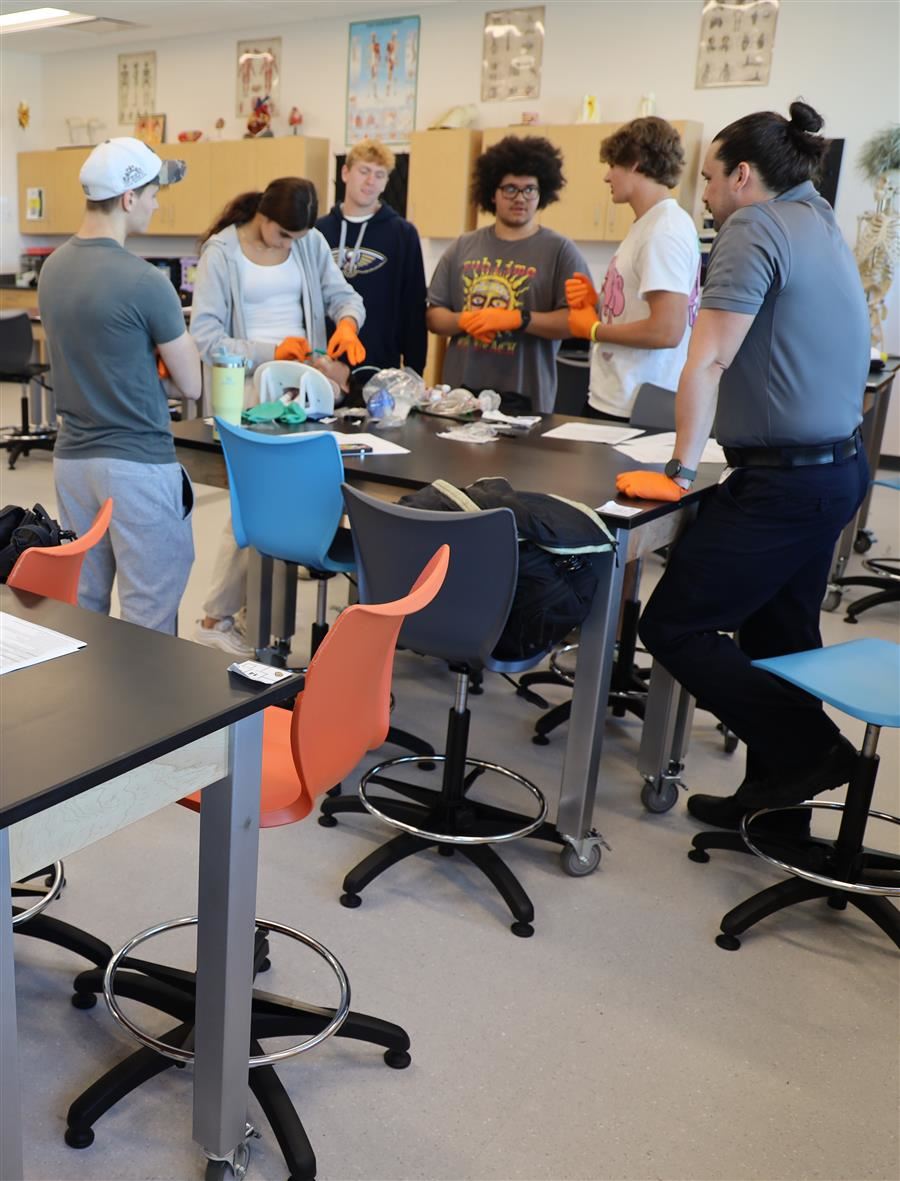  Students gather around a table in a science classroom to learn EMT skills.